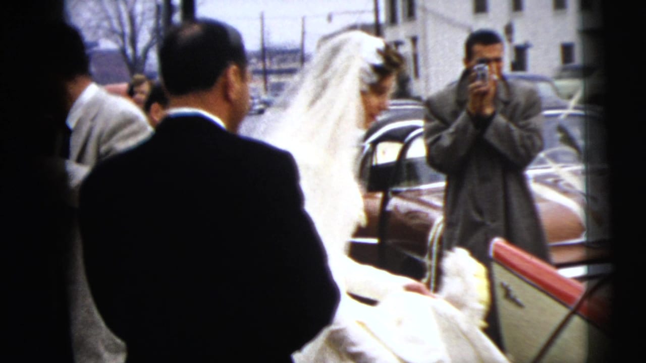 The bride nears a waiting car as suited guests watch and one man photographs the scene with a handheld camera.