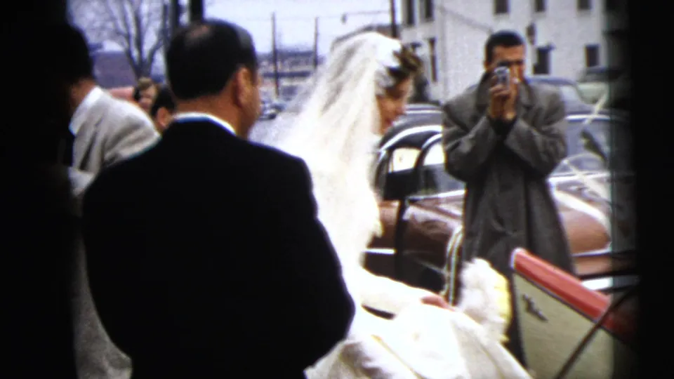 The bride nears a waiting car as suited guests watch and one man photographs the scene with a handheld camera.