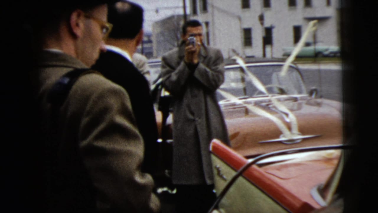 A photographer in a long coat faces the wedding party near a red-and-white convertible during the 1962 Moline curbside wedding scene.