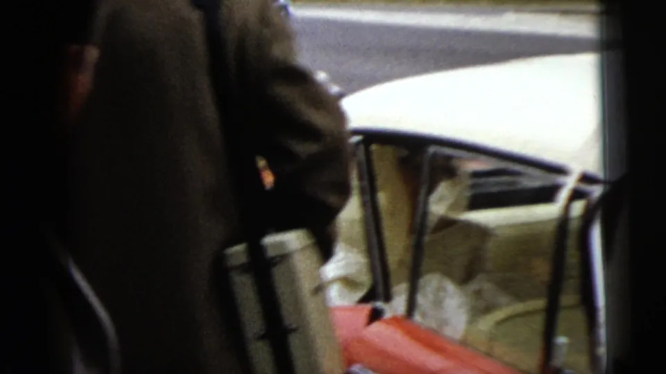 The bride appears inside or entering the open passenger side of the convertible while guests and the wedding party remain close to the curb.