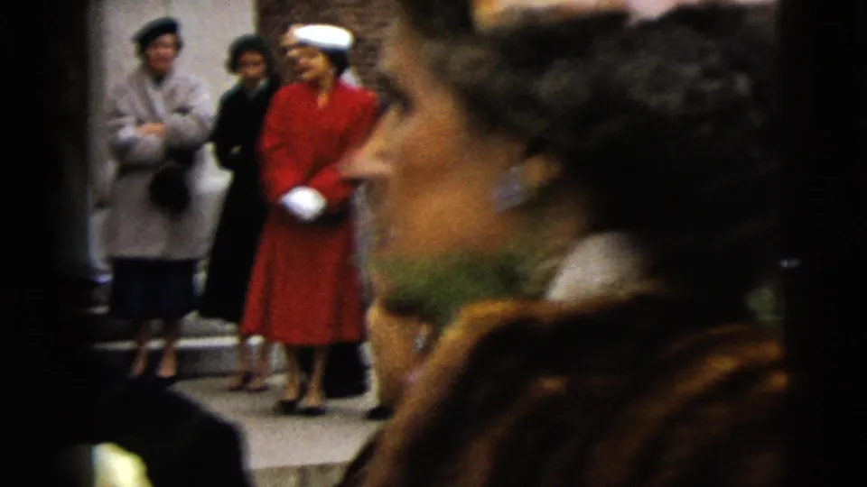 Women in period coats, including one in a red coat, watch from the sidewalk as the wedding departure continues in Moline, Illinois.