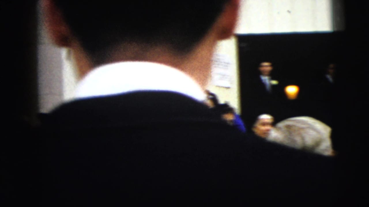 Back of a man in a dark suit frames people gathered near a building entrance.