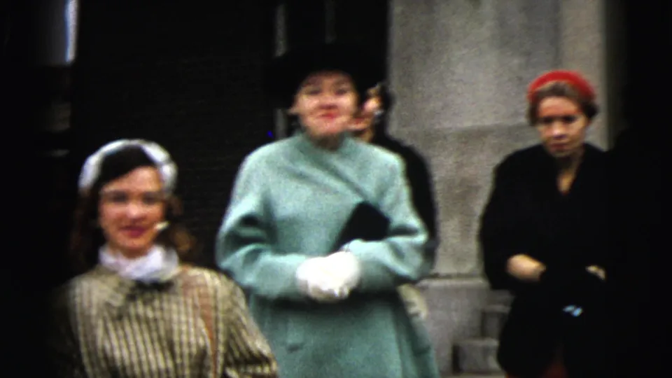 1962 Moline street frame showing women in formal coats and hats gathered by stone columns outside a building.