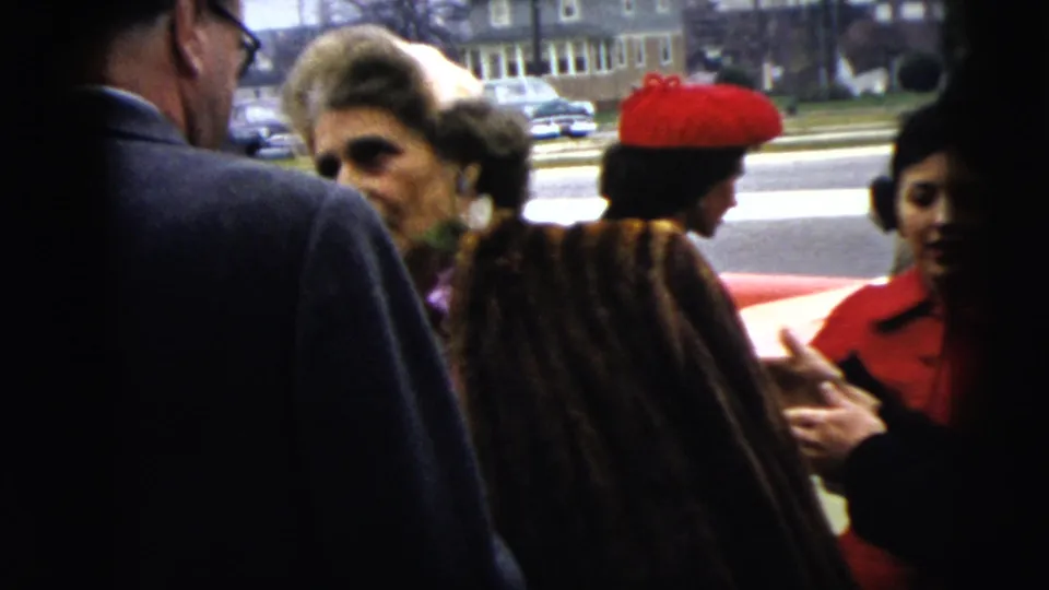 1962 Moline home-movie frame of adults talking on a sidewalk, with fur coats, hats, and a street with parked cars behind them.