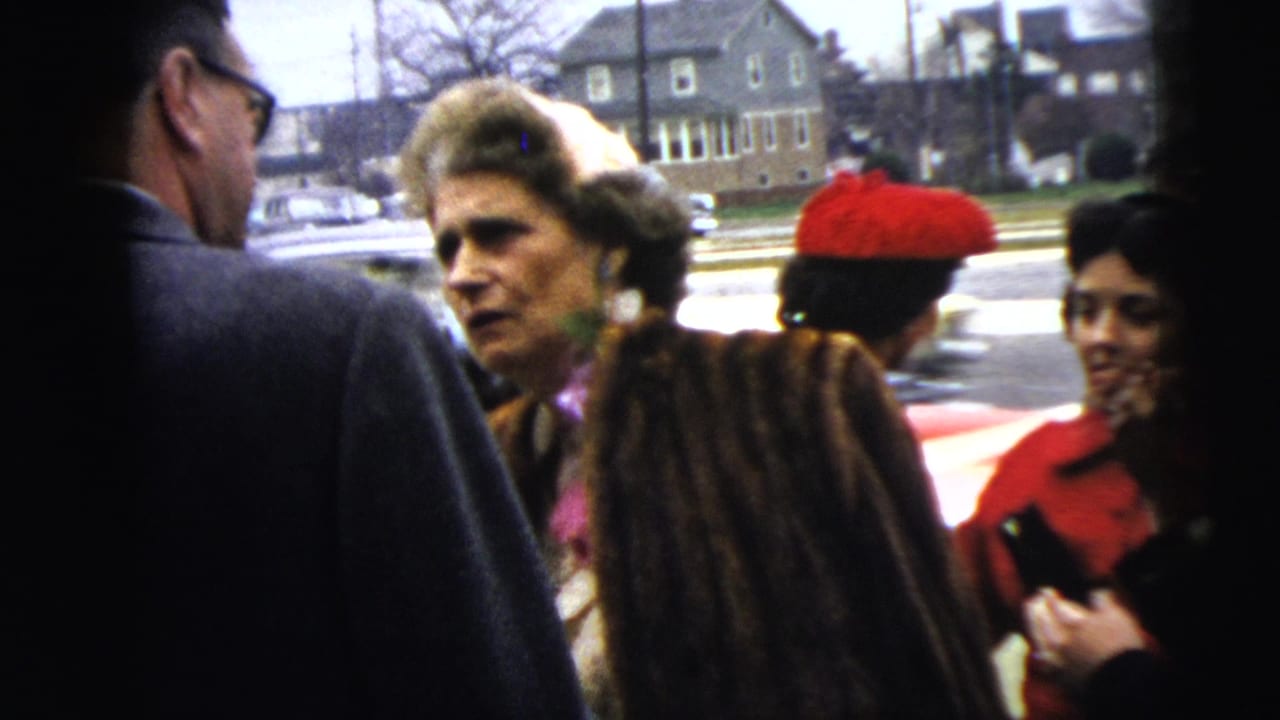 Adults in coats converse outdoors near a street with houses and parked cars behind them.