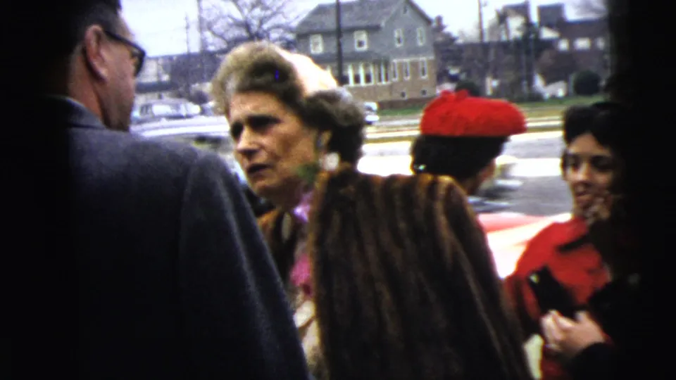 In Moline, Illinois in 1962, adults in period coats and hats stand close together during a sidewalk conversation near parked cars and homes.