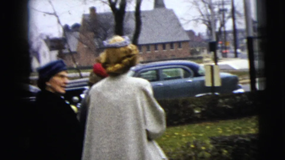 In the 1962 Moline footage, a woman in a white coat and patterned hat is seen from behind near a curb, with a blue mid-century car beyond her.