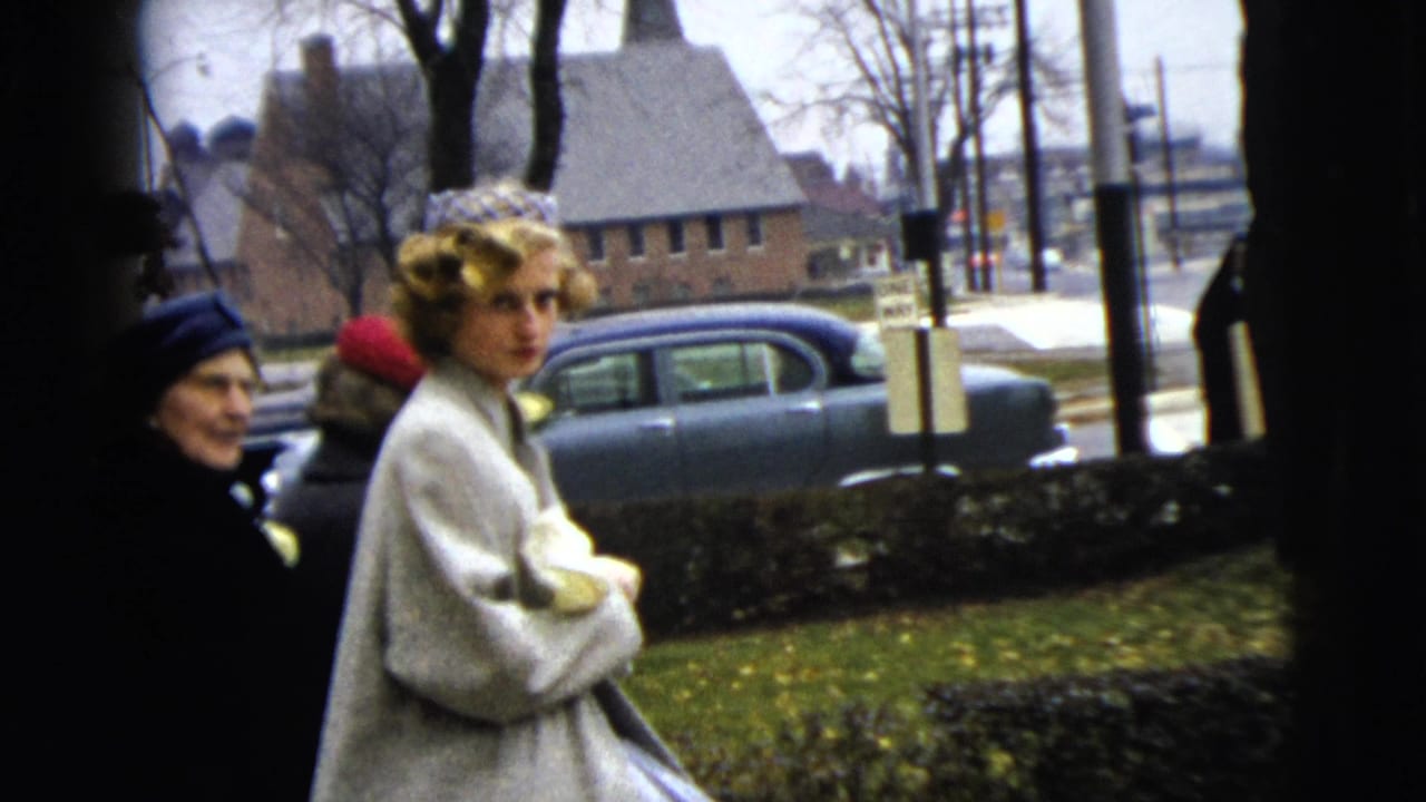 Woman in a light coat stands near other women on a sidewalk with parked cars behind her.