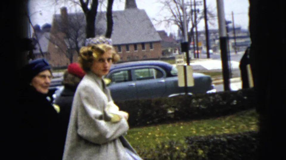 In a 1962 Moline street scene, a woman in a light dress coat faces the camera near other women and parked cars.