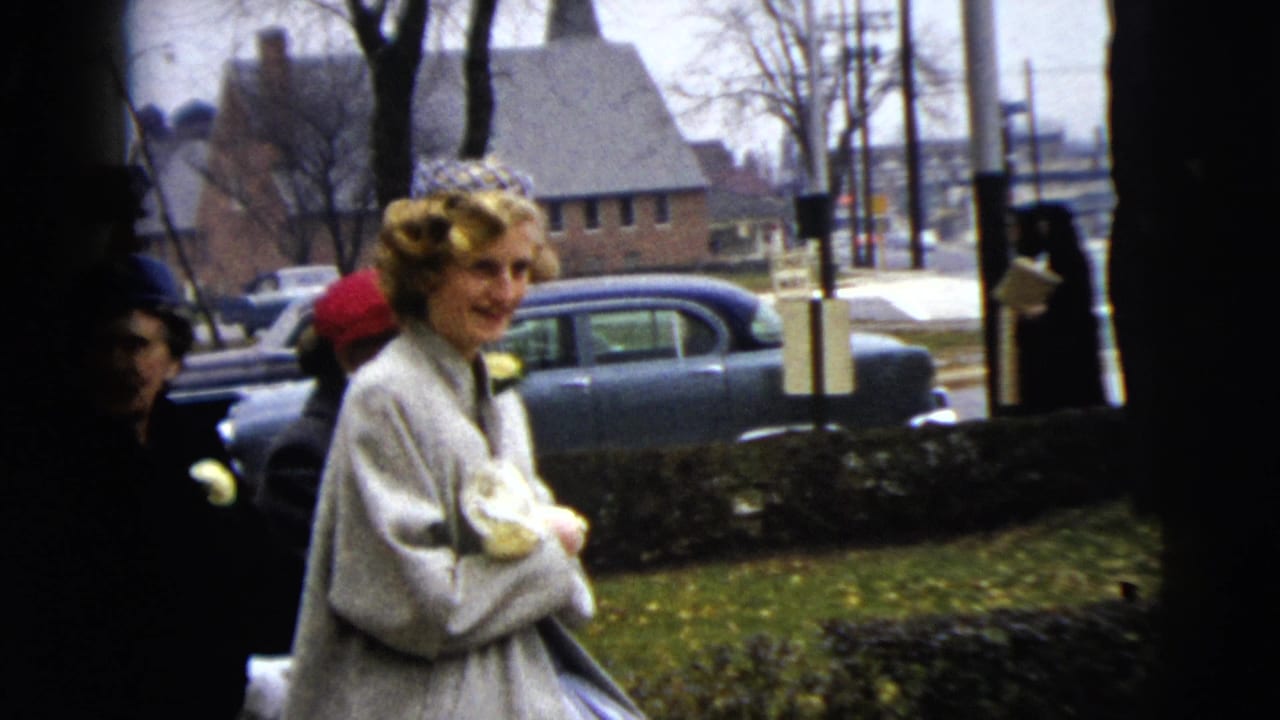 A woman in a light coat and decorative headband stands by a Moline sidewalk in 1962, with other women and cars behind her.