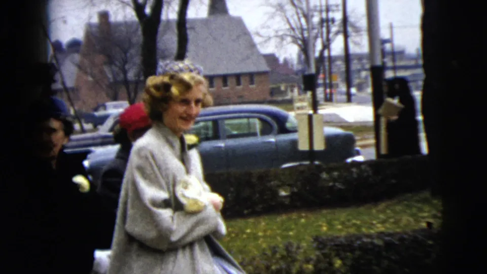 A woman in a light coat and decorative headband stands by a Moline sidewalk in 1962, with other women and cars behind her.