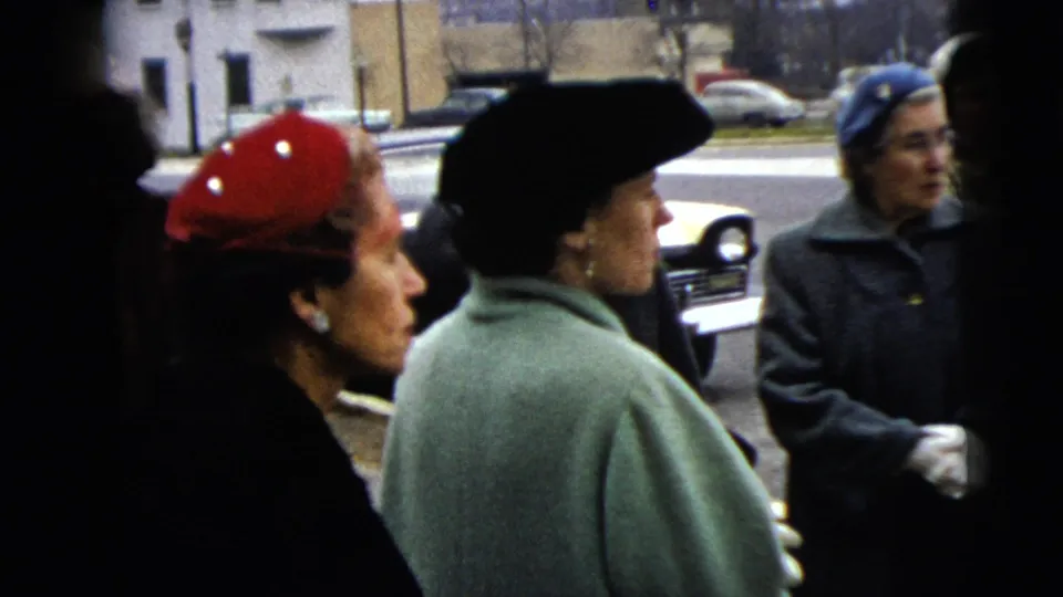 Women in red, black, and blue hats gather near the curb in a 1962 Moline street view with parked cars beyond them.