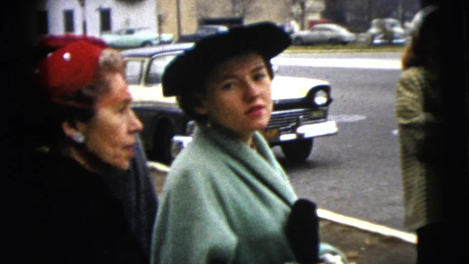 A woman in a pale coat and black hat faces the camera beside another woman in a red beret on a 1962 Moline street.