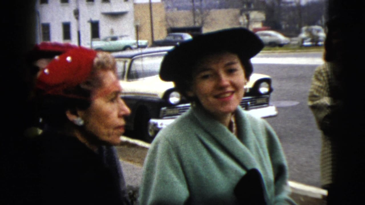 Two women in hats stand beside a street with parked cars behind them.