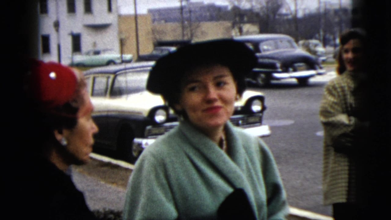 A young woman in a mint coat smiles while another woman faces her near parked cars.