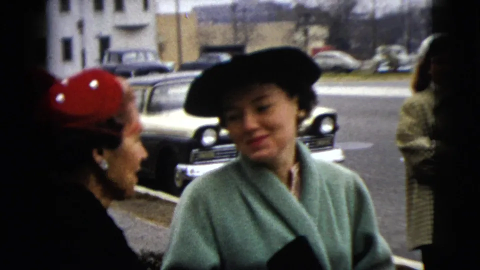 Two women appear to chat beside a Moline curb, with a two-tone car and low buildings visible in the background.