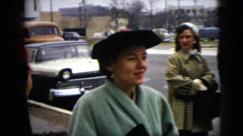 A woman in a mint coat and black hat looks away from the camera as another woman stands in the background near parked cars.