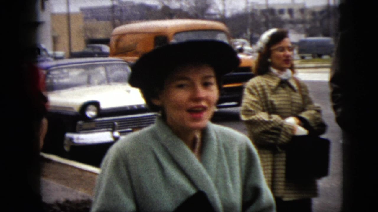 In supplied 1962 Moline home-movie footage, a woman in a black hat and mint coat faces the camera near early 1960s parked cars.