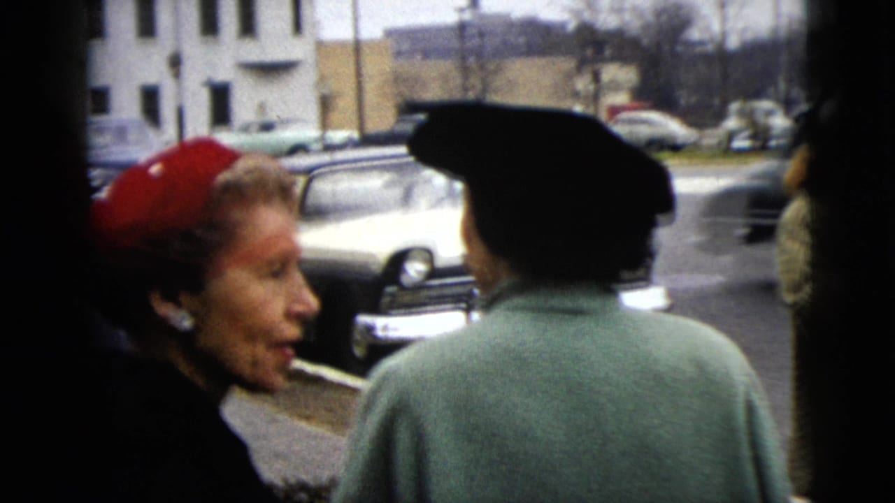 A woman in a red hat appears in profile beside another woman in a dark hat, with parked cars and buildings in the background.
