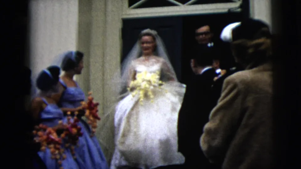 Bride in a white gown and veil stands at a Moline church entrance with blue-dressed bridesmaids and suited guests in 1962 home-movie footage.