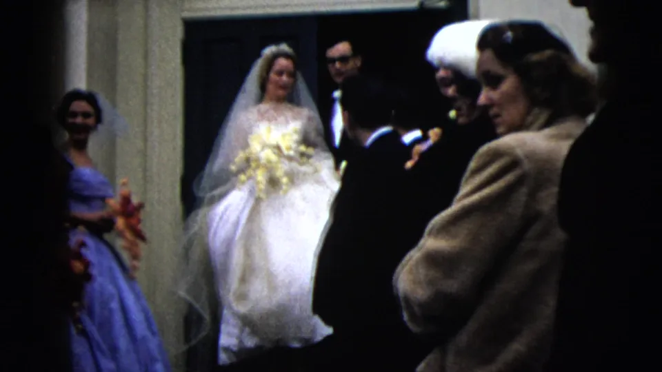 In 1962 Moline, Illinois, a bride in a white gown and veil stands near a doorway as bridesmaids and suited guests gather around her.