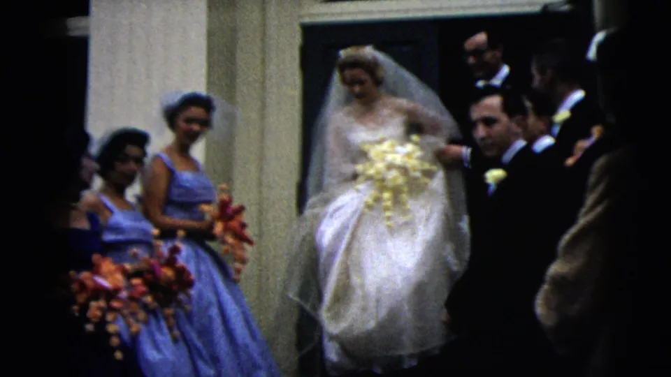 The bride continues forward past bridesmaids and men in dark suits, with bouquets and doorway columns visible in the 1962 Moline wedding clip.
