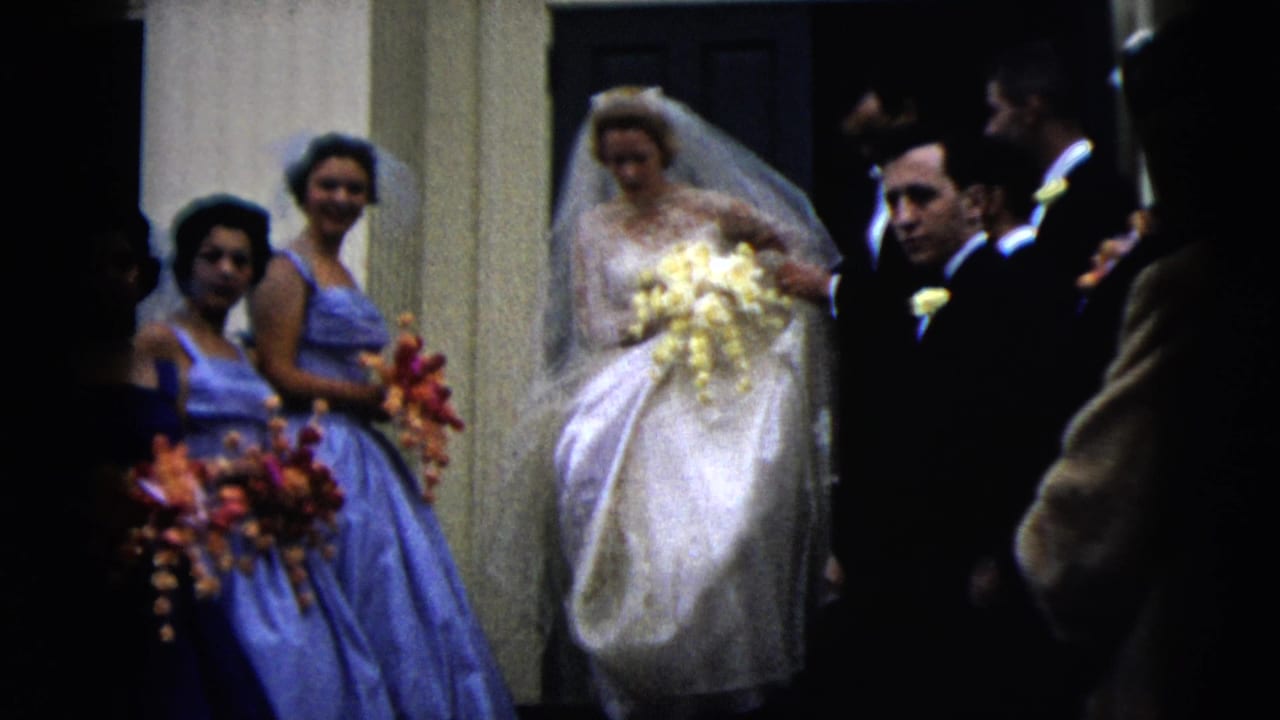 Bride and attendants stand near a doorway while guests and men in suits cluster around them.