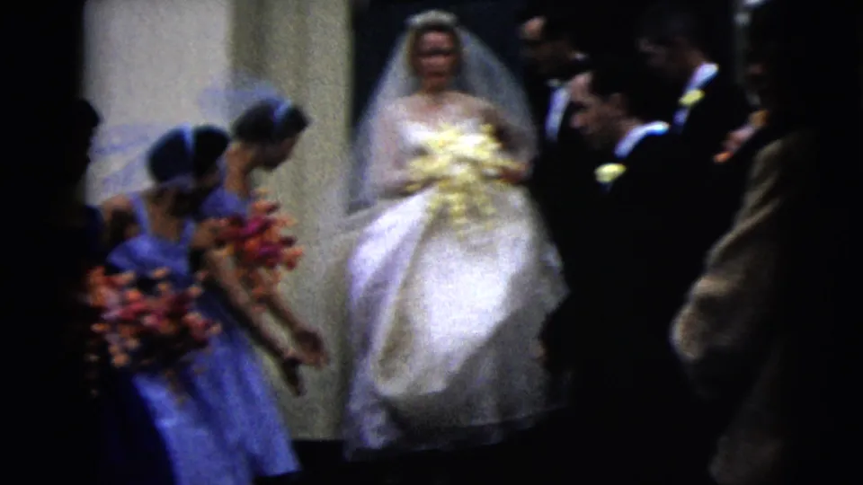 The bride moves from the doorway in 1962 home-movie footage, flanked by bridesmaids holding colorful floral bouquets.