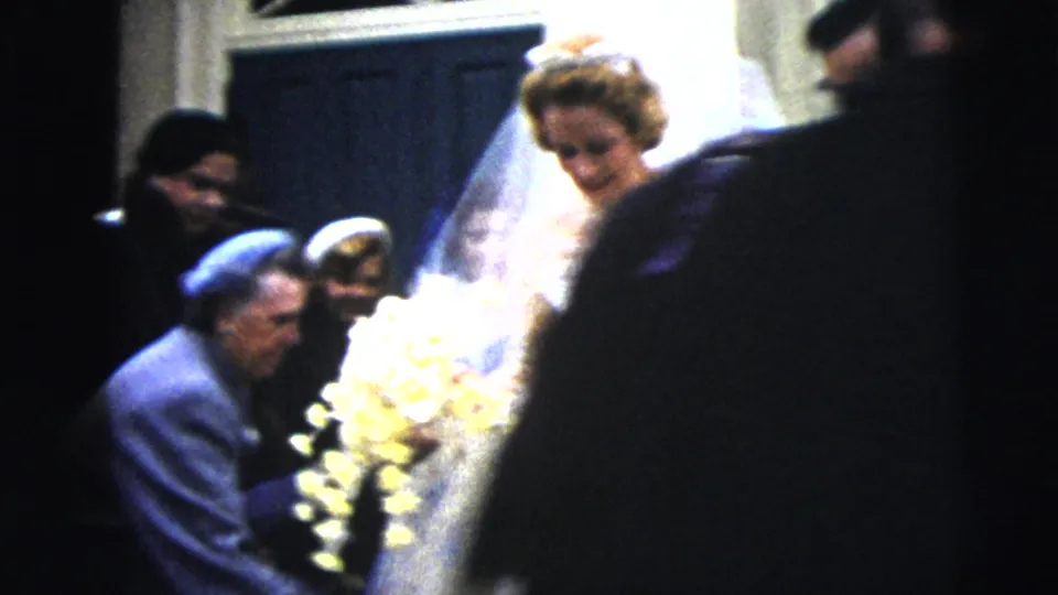 The bride pauses near guests at the church exit in Moline, Illinois, with her bouquet visible and the home-movie camera partly obscured by a foreground person.