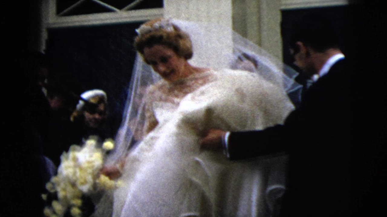 Bride in lace gown and veil descends stairs with a yellow bouquet as guests stand nearby.