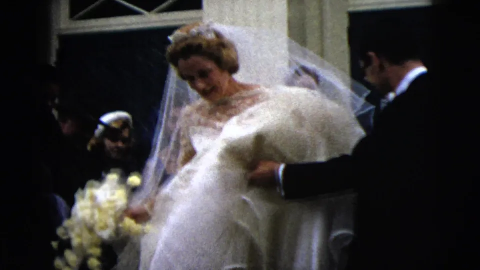 In 1962 Moline wedding footage, a bride in a lace gown and veil steps down stairs holding yellow flowers while guests watch.
