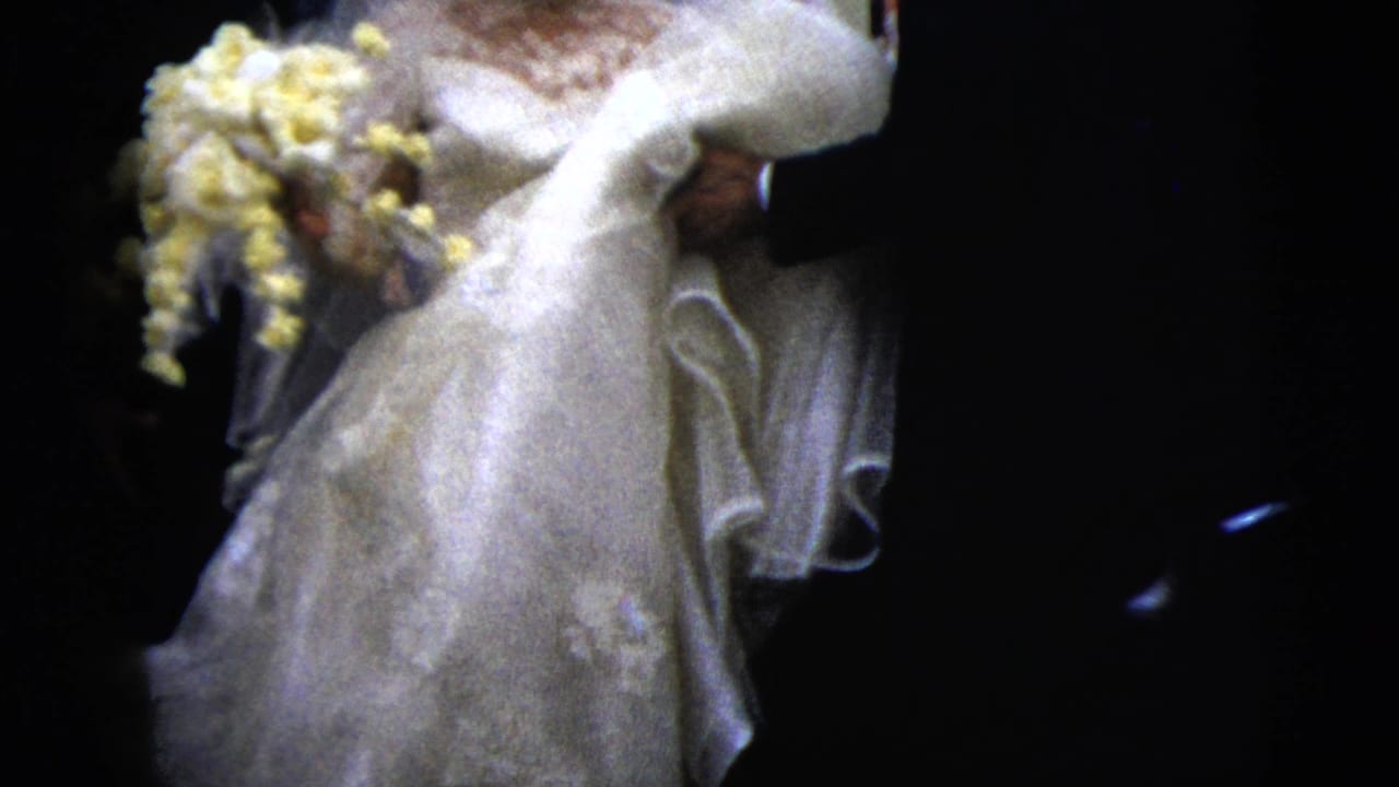 The bride's lace gown, veil, and yellow bouquet fill the frame as the groom walks beside her in this 1962 wedding home movie.