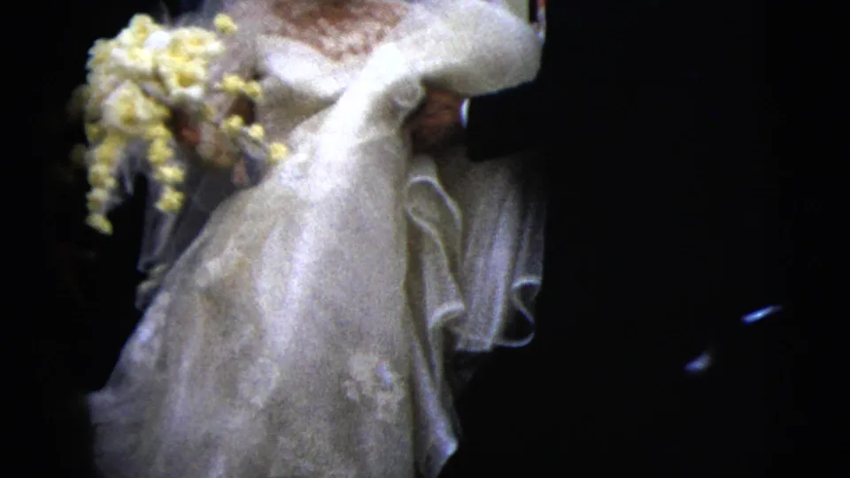 The bride's lace gown, veil, and yellow bouquet fill the frame as the groom walks beside her in this 1962 wedding home movie.