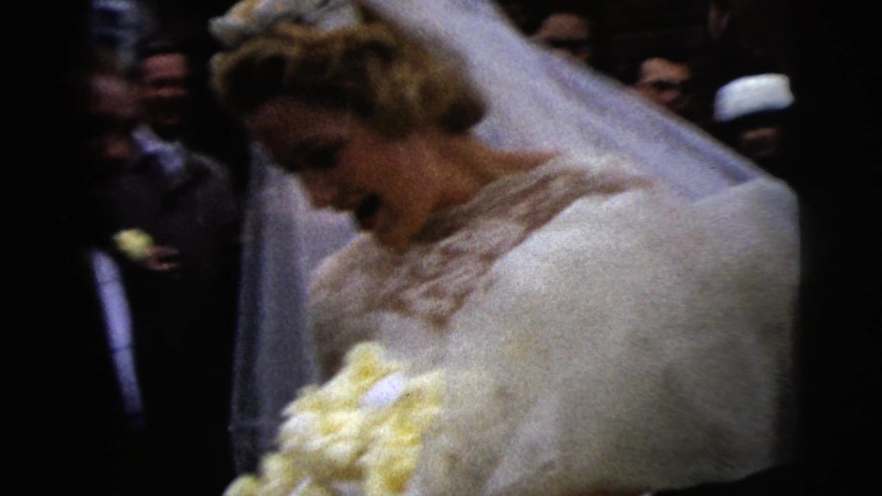 Bride in white dress and veil looks down while holding a yellow bouquet among wedding guests.
