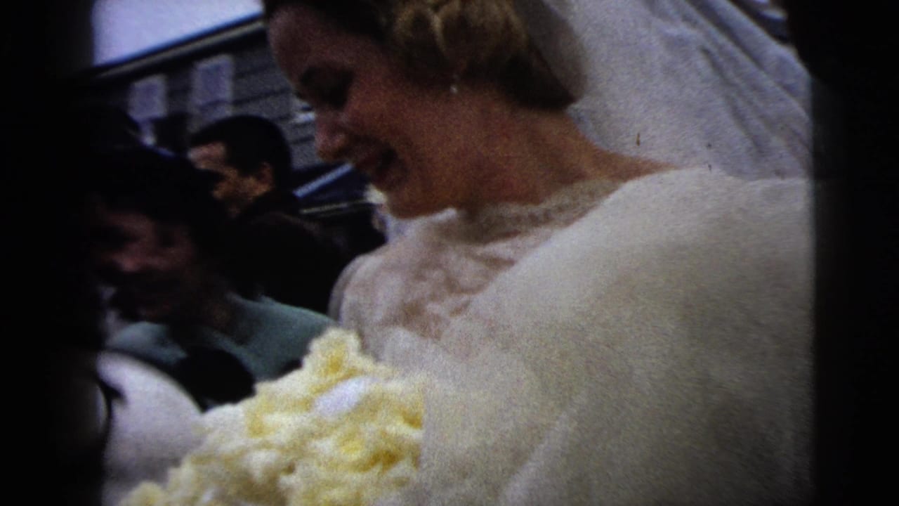 Smiling bride in veil carries a yellow bouquet beside guests in formal clothing.