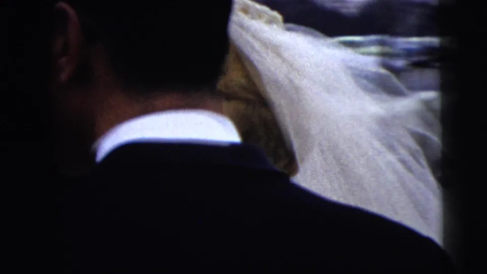 1962 Moline wedding home movie frame showing the bride's veil from behind as dark-suited guests crowd near parked cars.