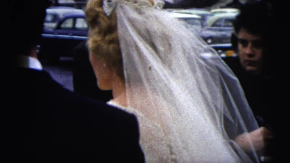 Bride in a white veil stands beside a dark-suited man during the 1962 Moline wedding departure, with bystanders and cars behind them.