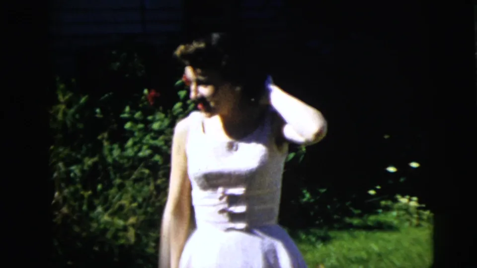 Adult woman in a white sleeveless dress stands in a sunlit Moline garden, with dark foliage and lawn around her.