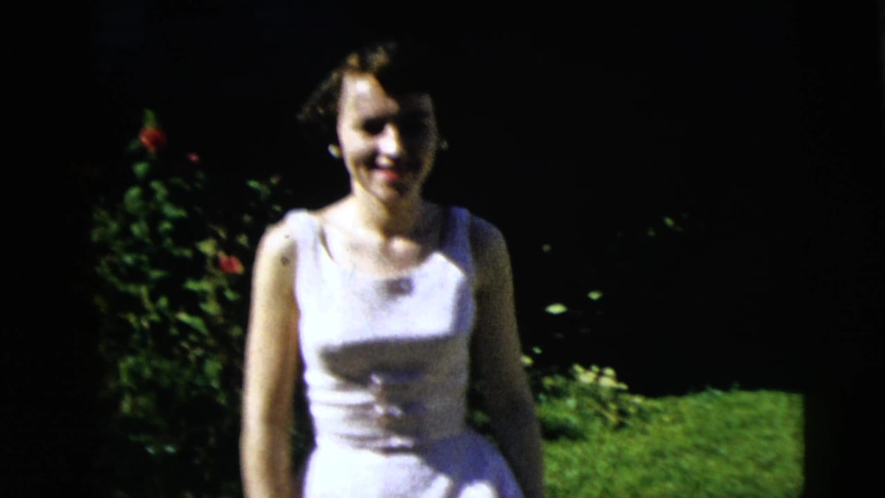 Young woman in a white dress smiles in a sunlit Moline yard, with red flowers and grass behind her.