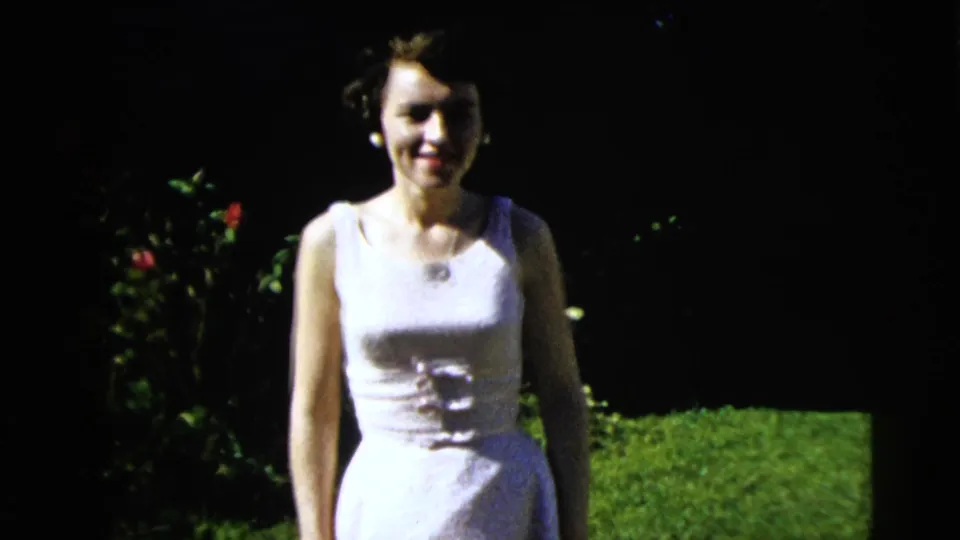Young woman in a white dress smiles in the yard, with lawn and rose bushes visible in Moline, Illinois home-movie footage.