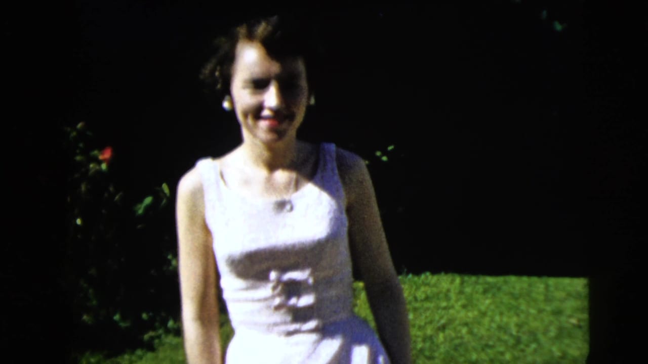 Young woman in a white sleeveless dress smiles while walking across a lawn.