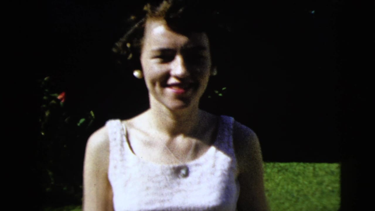 Smiling woman in a white dress stands closer to the camera against lawn and shrubs.