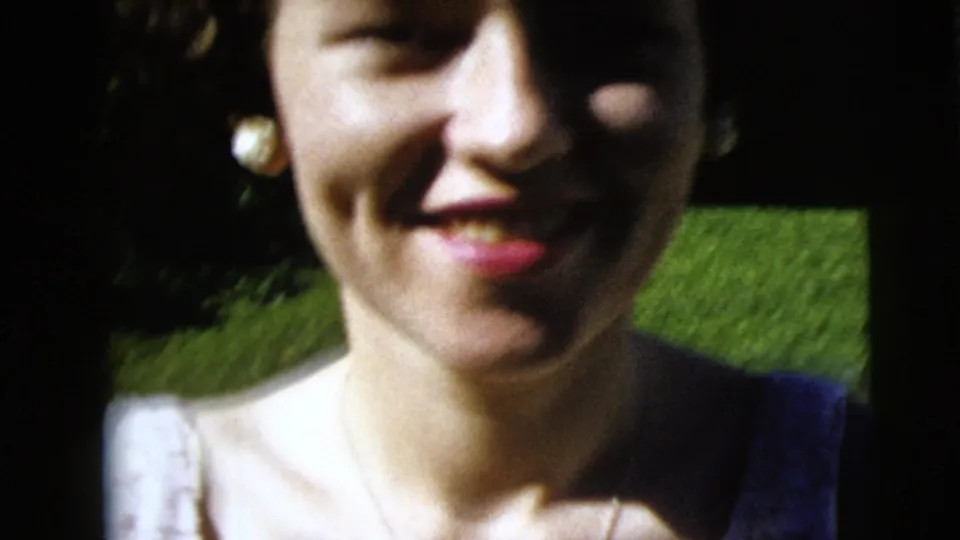 Close home-movie view of the woman's smile, pearl earrings, and white dress, with green lawn visible behind her.