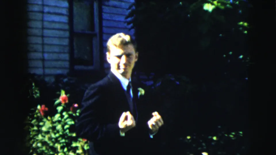 A young man in a dark suit and tie stands by flowering shrubs before a sided house in 1962 Moline home-movie footage.