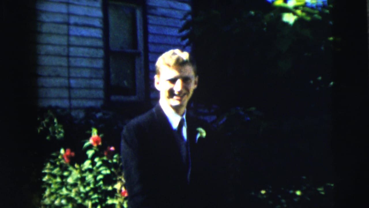 Young man in a dark suit stands outside near flowers and house siding.