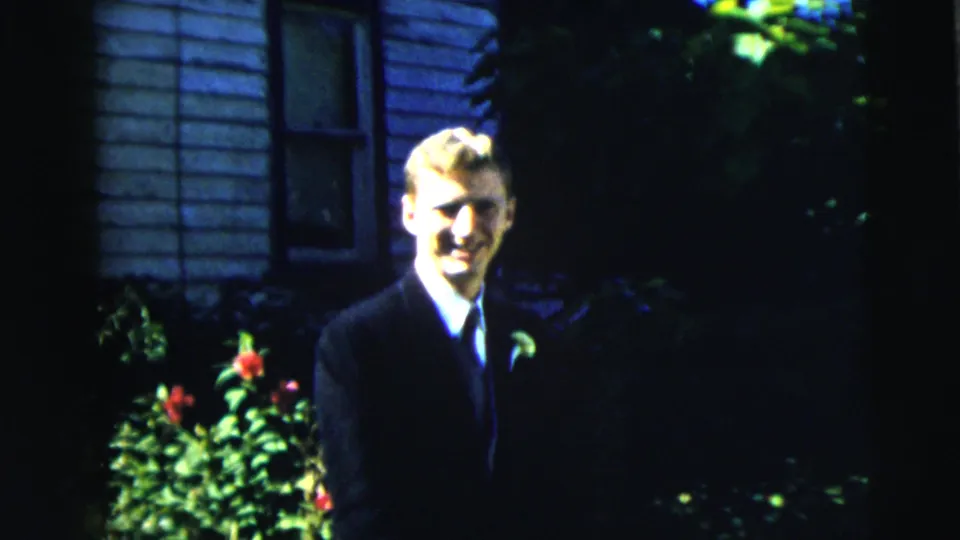 Young man in a dark suit and light tie stands outside a Moline, Illinois house in 1962, with flowers and foliage nearby.