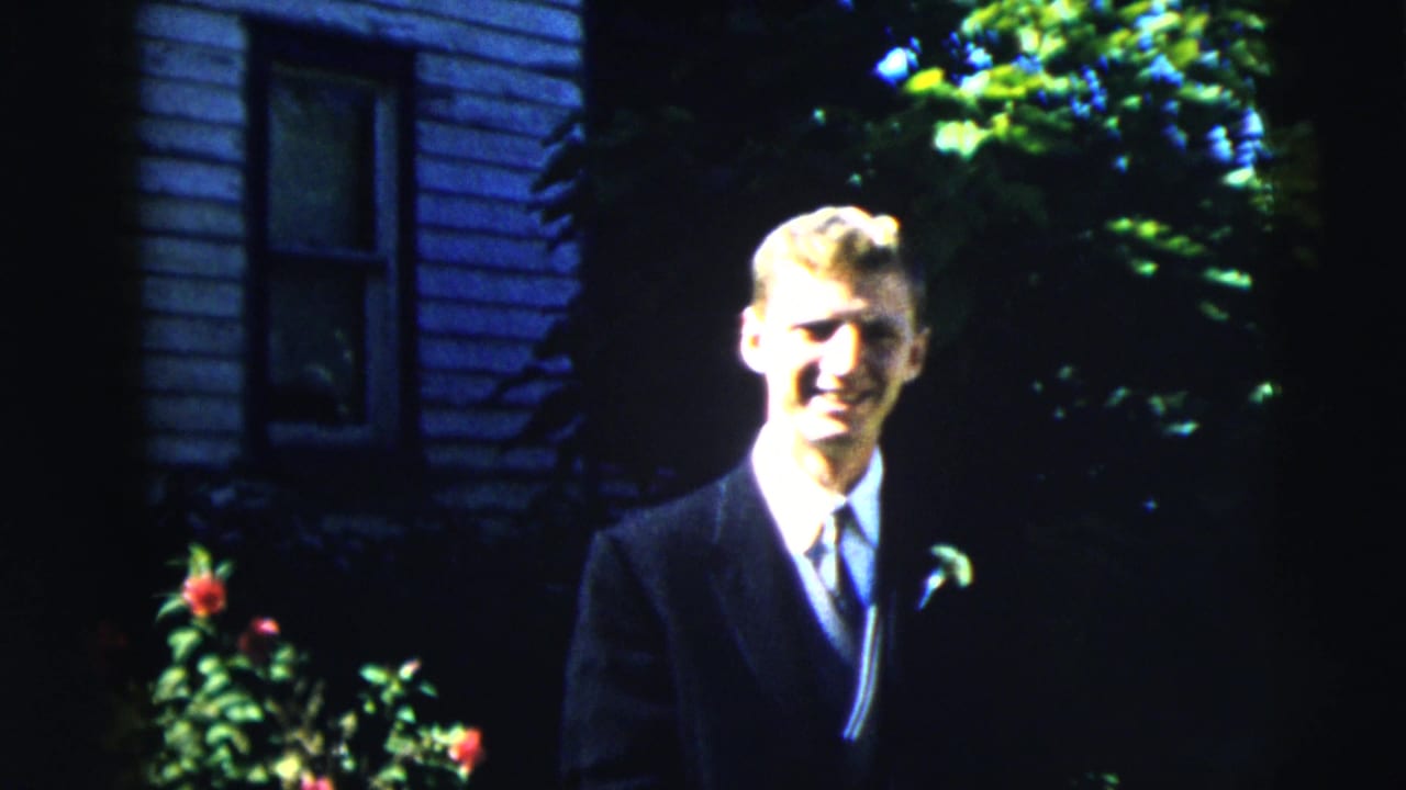 Young man in suit smiles toward the camera beside leafy plants.