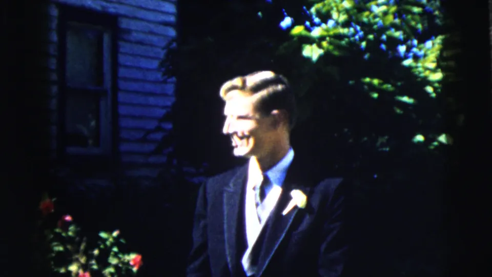 The suited young man turns in profile and smiles outdoors in Moline, Illinois, with garden flowers and house siding visible.