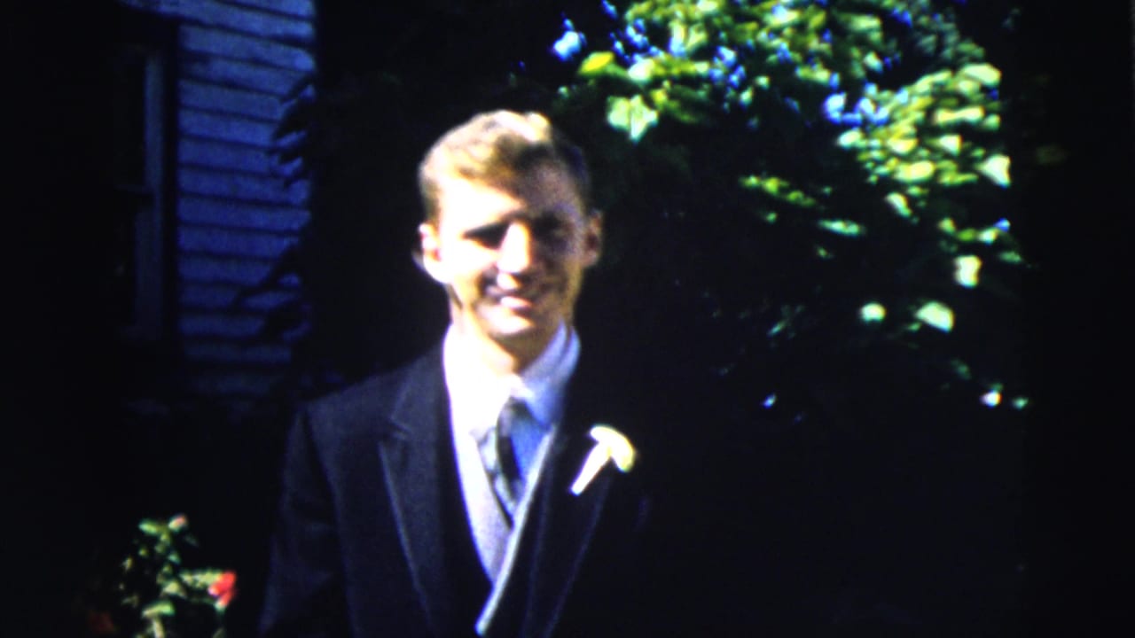 Young man in a dark suit with a boutonniere standing outdoors near house siding and greenery.