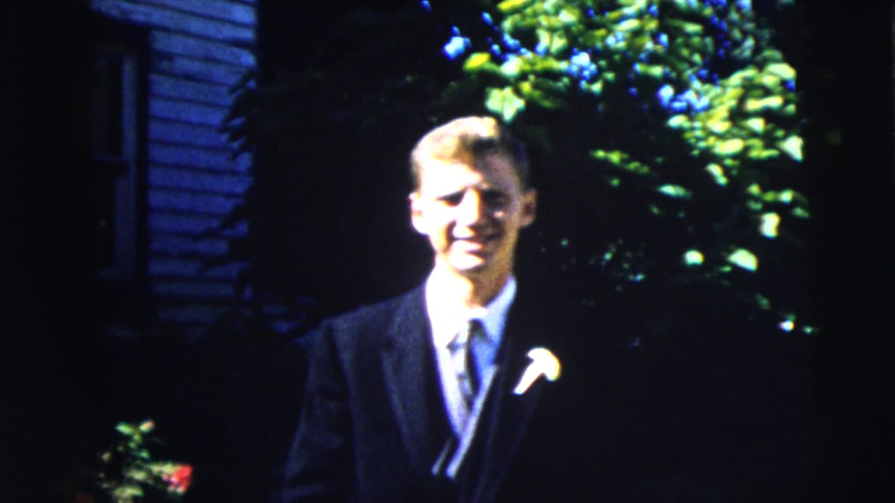 In 1962 Moline, a young man in a dark suit and boutonniere stands in strong sunlight before a leafy backdrop.